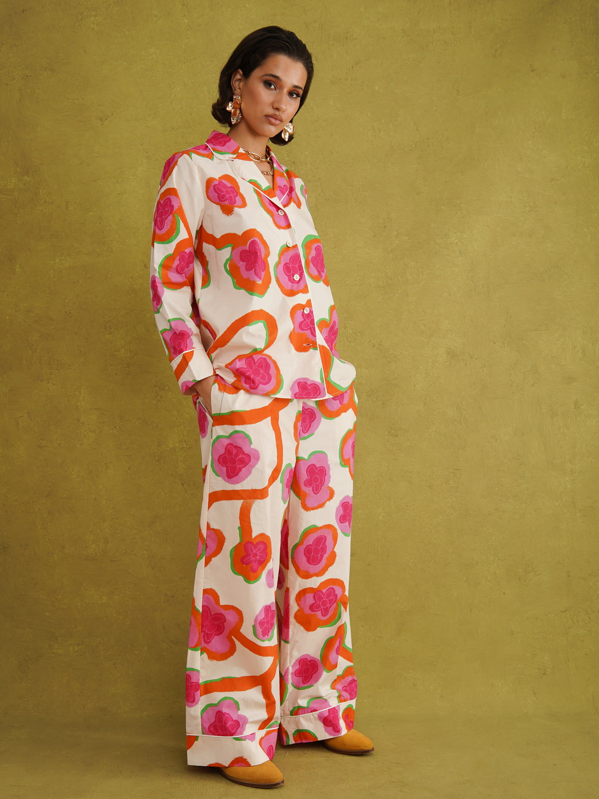 Clara Maria wearing the Jackie Pyjama Set in size M-L with mustard flats: a loose cotton shirt and wide-leg trousers in a cream print of hot pink flowers ringed in orange and green